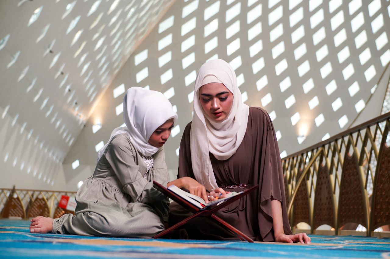 A mother and daughter wearing hijabs reading the Quran in a beautifully lit mosque interior.