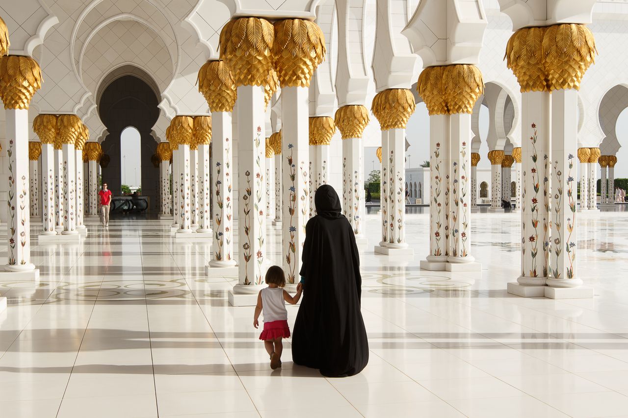 An adult and a child walk together between the majestic pillars of a grand mosque.
