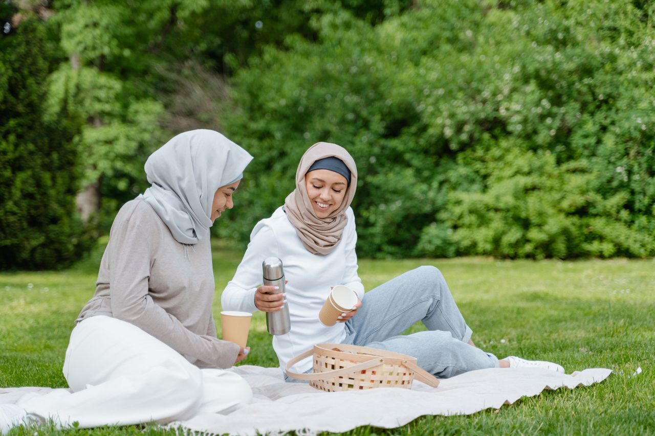 Two women wearing hijabs having a picnic in the park with paper cups and a vacuum flask.
